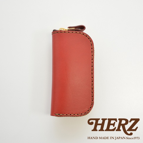HERZ 品牌標誌，下方有 HAND MADE IN JAPAN Since1973 字樣，圖片主體為紅色皮革製的橢圓形拉鍊鑰匙包，有四鉤，採用手工縫線。