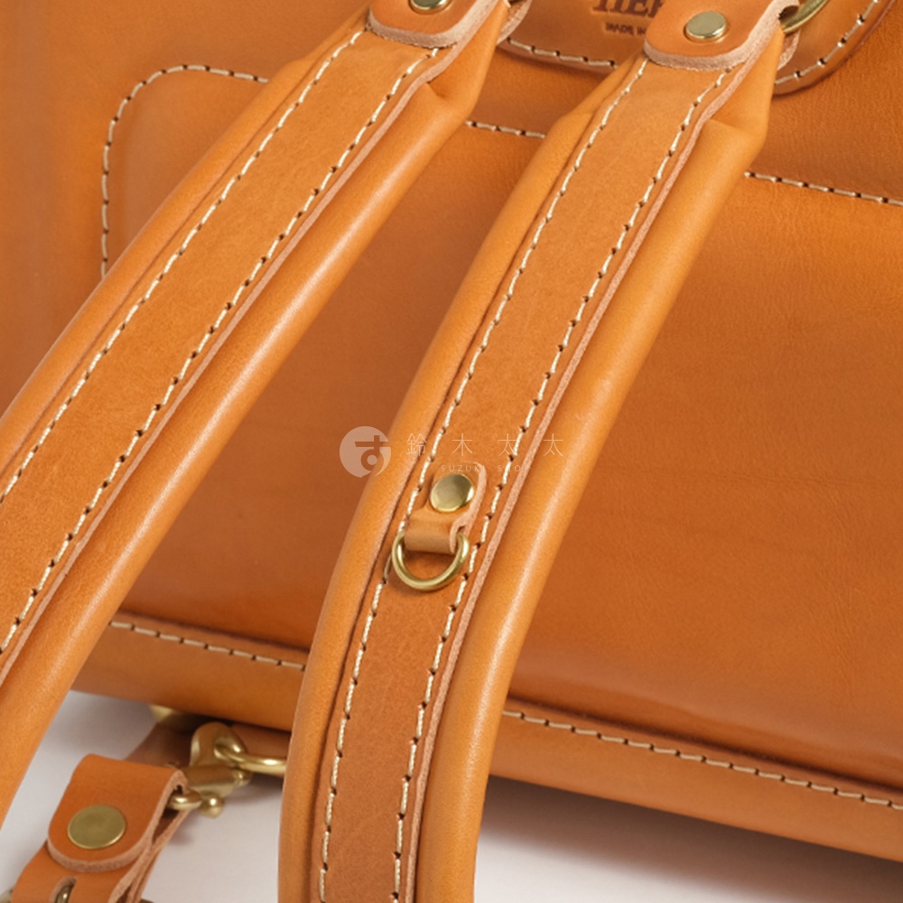 職人手作皮革橫式書包的揹帶特寫，皮革顏色為暖棕色，縫線清晰，金屬扣環精緻。