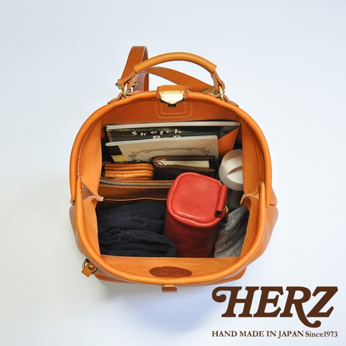 一個打開的橘棕色皮革後背包，內部裝有多本筆記本、一個紅色皮革小包、黑色衣物和一個白色物品，背包頂部有提把和金屬扣。