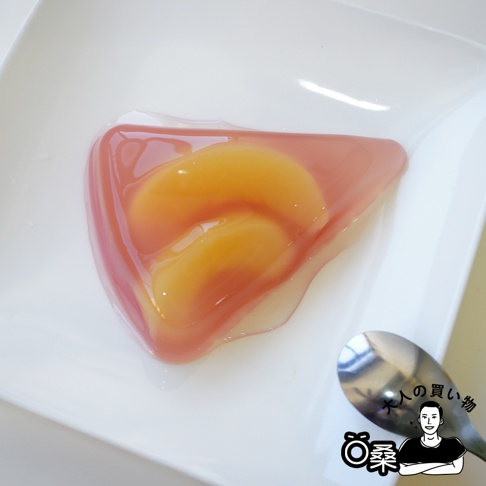 粉紅色果凍中包裹著兩片金黃色桃子果肉，呈三角形，置於白色盤中，盤邊有一支金屬湯匙。