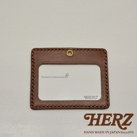 職人手作皮革橫式證件識別夾，咖啡色皮革，邊緣縫線精緻，包含一個用於懸掛的金色圓孔及透明證件視窗，內有「The Heart of Craftsmanship」文字與小圖案，右下角印有「HERZ」標誌。