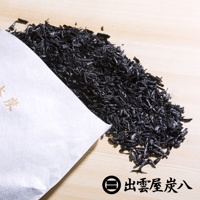 一堆細碎的黑色木炭散落在淺色木紋背景上，部分木炭從白色紙袋中傾倒而出，紙袋上有金色日文字樣。