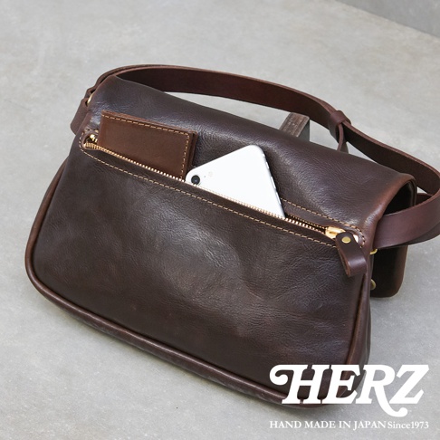 職人手作皮革斜背包，深棕色，有拉鍊主袋和外袋，展示手機和卡夾，皮革質感。