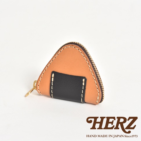 日本HERZ手工皮革零錢包，外觀設計為可愛的三角飯糰造型，採用Latigo軟皮革製作，搭配對比鮮明的黑底與淺棕色皮革，邊緣有精緻的手縫線細節，並配有拉鍊開口，右下角印有HERZ品牌標誌與手工產地說明。