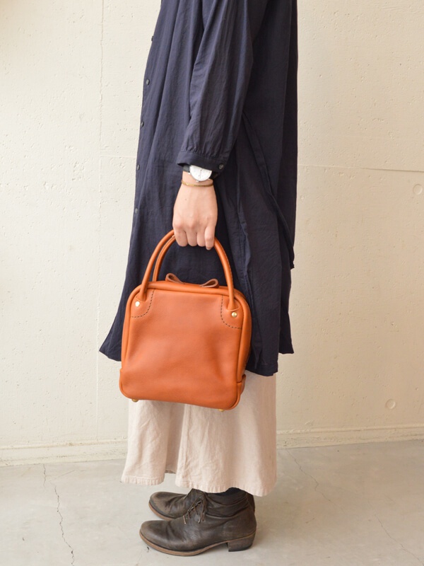 職人手作皮革Organ方形托特包，採用溫暖的橘棕色皮革，包身線條簡潔，頂部有兩個短提把，皮革表面帶有細緻紋理，四個角以金屬釘固定並縫線裝飾，整體呈現質感。