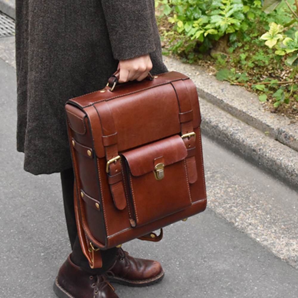 棕色皮革學院風後背包，配有翻蓋和金色搭扣，側面有皮帶和金屬釘飾，提把和部分肩帶可見。