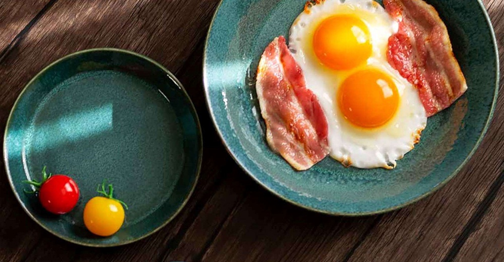 兩個綠松石色調的陶瓷盤子，一個盤子裡放著煎蛋和培根，另一個盤子裡放著紅色和黃色的小番茄，盤子放在深棕色木桌上。