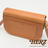 職人手作皮革經典馬鞍包，採用天然皮革材質，呈現溫潤的棕褐色。包款設計簡約，帶有明顯的縫線細節，正面有一個方形的貼袋，上面壓印著「HERZ」品牌標誌。