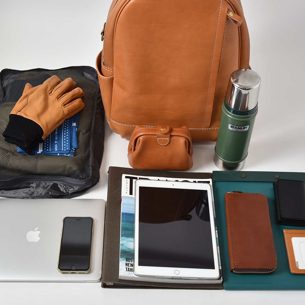 職人手作皮革多功能後背包 R-21，搭配棕色皮革手套、綠色保溫瓶、平板電腦、手機、皮夾及收納袋。