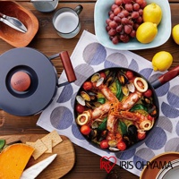 深灰色不沾平底鍋，鍋蓋配有木紋把手，鍋內盛有海鮮飯，配有蝦、蛤蜊、番茄和青豆。旁邊有葡萄、檸檬、起司和餅乾。