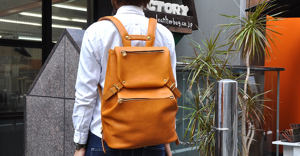 棕褐色皮革後背包，具有兩個前置拉鍊口袋和可調節肩帶，風格簡約。