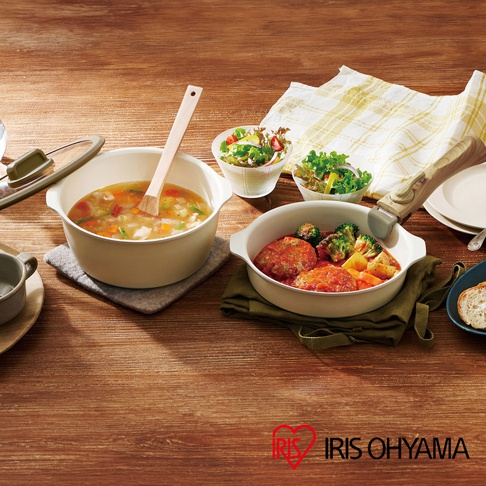 米白色陶瓷不沾鍋中盛有蔬菜湯，一旁另有一鍋裝有漢堡排、花椰菜和紅蘿蔔塊。鍋具皆有木紋把手，搭配兩小碗新鮮沙拉，整體擺設於木質桌面。