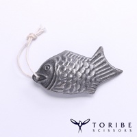 銀灰色金屬製成的小鯛魚造型，帶有一條米白色細繩，繩子末端打結。鯛魚表面刻有魚鱗紋路，尾部呈扇形。