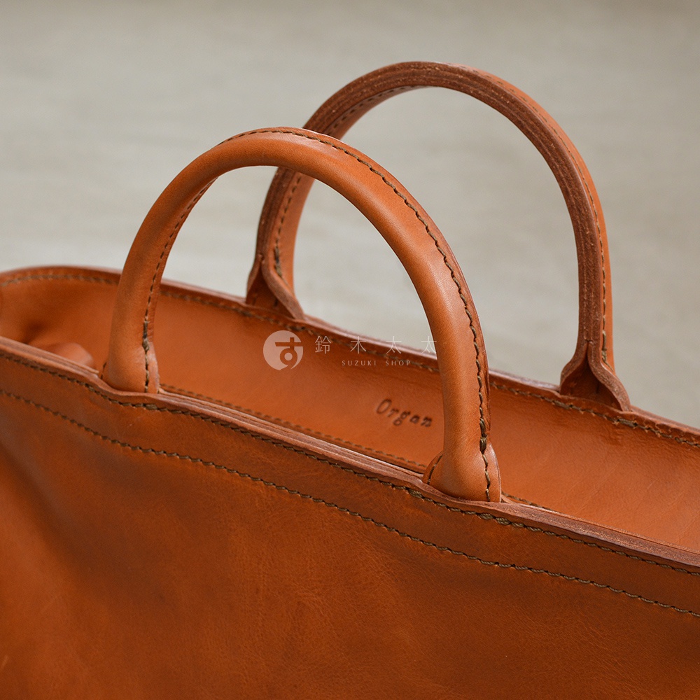 日本HERZ職人手作皮革Organ兩用拉鍊托特包，特寫焦糖色皮革的提把和縫線細節，包身右側可見品牌標誌及Organ字樣