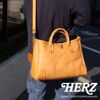 一位人士斜肩背著一個天然色皮革製成的大容量托特包，包包有兩個提把，並配有可拆卸的長背帶，包身側面有加固的皮革設計。