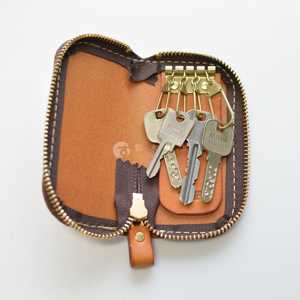 職人手作皮革橢圓形拉鍊鑰匙包，內部有四個金屬掛鉤，上面掛著幾把鑰匙，拉鍊為金色，皮革為棕色。