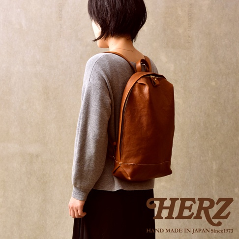 一位短髮女性背著日本HERZ手工製作的棕色簡約皮革後背包，該包款採用柔軟的義大利Eufrate皮革，側邊呈現獨特褶皺工藝，整體造型俐落且具備極簡美感。