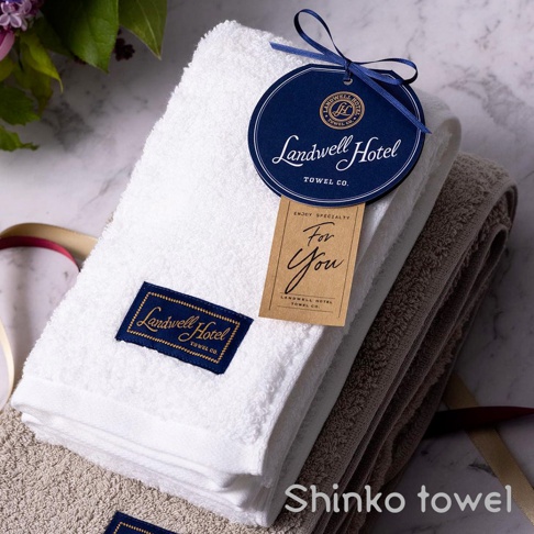 Landwell Hotel 泉州精繡長纖匹馬棉毛巾，展示白色和灰褐色兩款。白色毛巾上有藍色燙金標籤，並繫著圓形品牌吊牌及「For You」小卡。