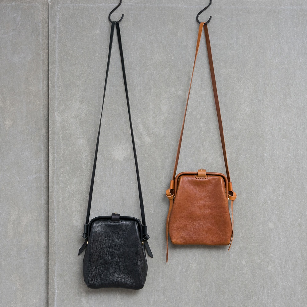 黑色與棕色兩款職人手作皮革Organ迷你杜勒斯側背包，以柔軟皮革製成，設有金屬扣環和可調節的細長斜背帶，垂掛於灰色水泥牆上。