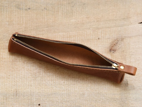 職人手作皮革Organ三角收納筆袋，顏色為棕褐色，材質為皮革，配有金色拉鍊和皮革拉環，置於木質背景上。