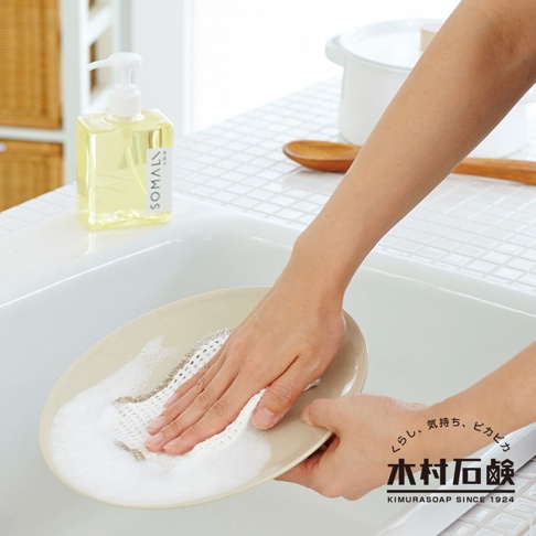 一個透明方形按壓瓶裝有淡黃色液體，標籤上印有「SOMALI」字樣。旁邊是一隻手正在用白色網狀海綿清洗盤子，盤子上有泡沫。