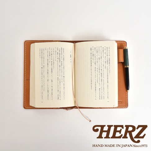 一本打開的皮革手帳，內頁有日文文字，旁放一支黑色鋼筆，手帳右下方有HERZ的Logo。