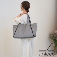 一位女士肩背一款黑色與白色細點刺子織紋理的托特包，包身寬大，有深色滾邊與提把，包口中央有鈕扣。