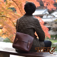 日本HERZ職人手作皮革雙扣旅行郵差包，以Latigo軟皮革製作，深棕色包款配有雙插釦，背對鏡頭坐在木凳上，背景是秋季楓紅與日式建築