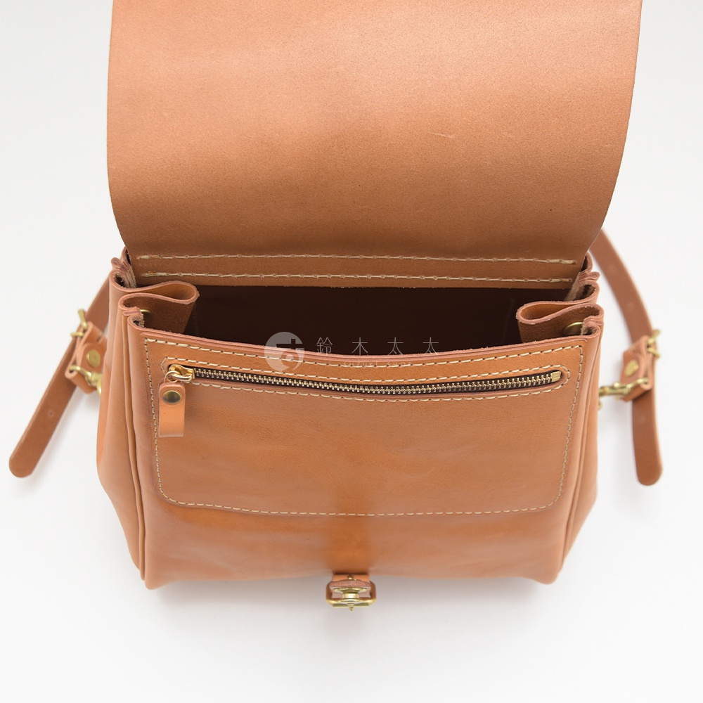 職人手作皮革屋瓦風後背包，淺棕色，翻蓋設計，外部有拉鍊口袋與金屬旋轉鎖扣，可見皮革紋理與縫線細節。