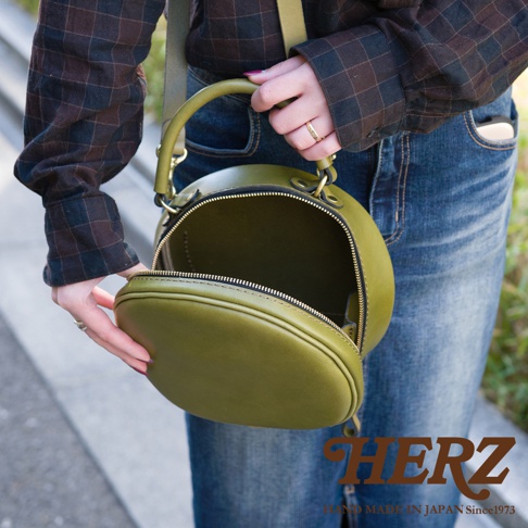 日本HERZ品牌復古圓形皮革包，以綠色軟皮革製成，可手提或斜背，包包開口露出內部，下方有HERZ品牌標誌。