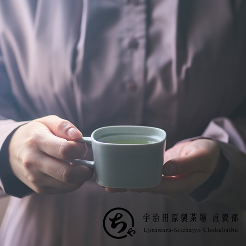 一位身穿淺紫色襯衫的人雙手捧著一個簡約質感的灰色美濃燒馬克杯，杯中盛裝著綠茶，畫面右下角標有宇治田原製茶場直賣部的品牌標誌與文字。