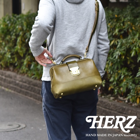 一位穿著灰色連帽衫的男士背著日本HERZ手作橄欖綠皮革口金單肩包，包款採用經典醫生包設計配有金屬釦飾與提把，背帶可拆卸並能調整長度，整體呈現精緻職人風格。