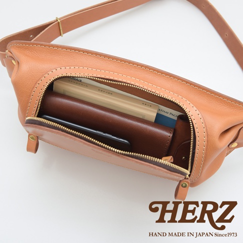 HERZ 品牌皮革拉鍊旅行斜背包，內部有一個皮夾和手機，上方有一本書，標示為HAND MADE IN JAPAN Since1973