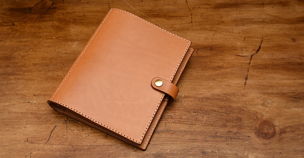 職人手作皮革A5手帳/記事本書套，淺棕色皮革，配有同色皮革束帶與金色按扣，側邊縫線精緻，置於木紋桌面。