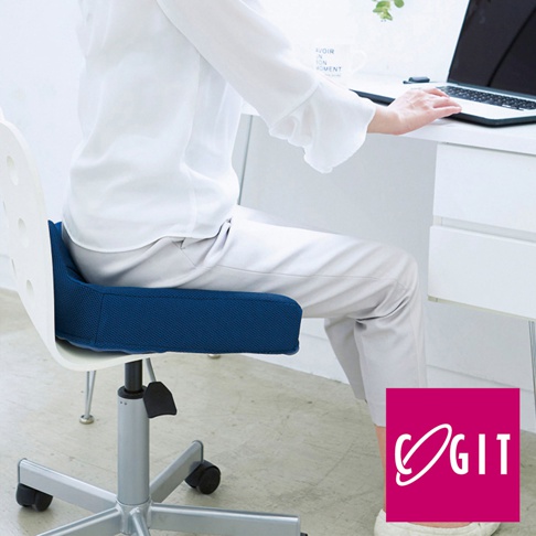 深藍色網狀材質的W型機能紓壓坐墊，置於白色辦公椅上，一位身穿白色上衣、米色長褲的人士正坐於其上，前方為白色桌面與筆記型電腦。
