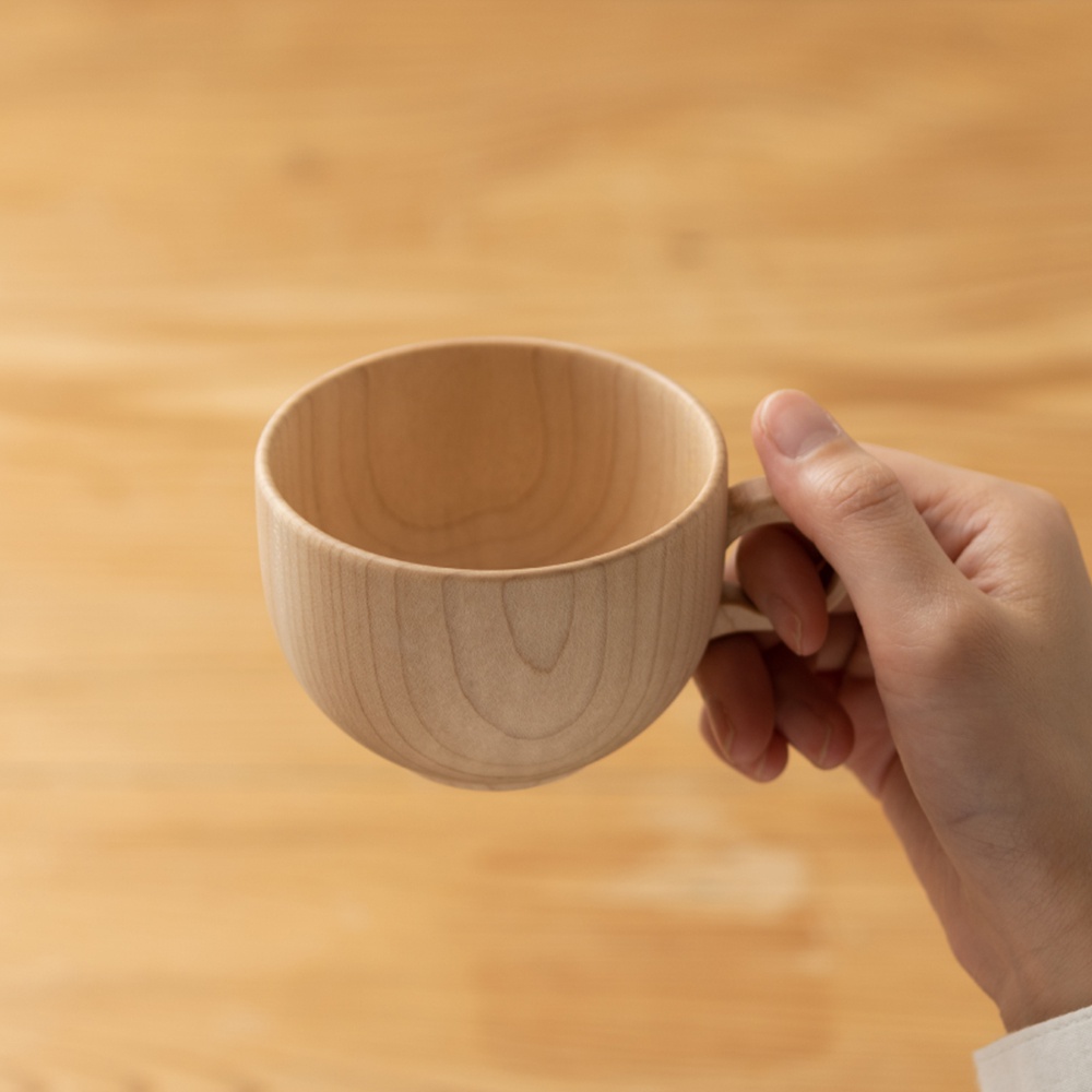 一只手正握著一個淺色楓木茶杯，杯子呈現自然的木紋，帶有圓潤的把手，背景為木質桌面。