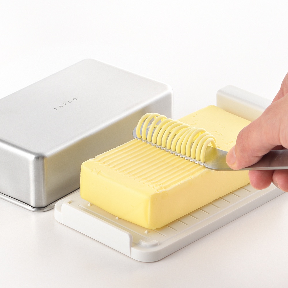 日製奶油盒，銀色金屬盒蓋印有「EATCO」字樣，搭配白色奶油盒，盒內有一塊黃色奶油，正由一支銀色金屬奶油刀刮出螺旋狀奶油絲。