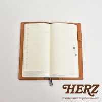 淺棕色皮革製成的旅行記事本書套，打開展示內頁，左頁為日曆格式，右頁為方格筆記頁，書套邊緣有縫線裝飾，下方有品牌標誌。