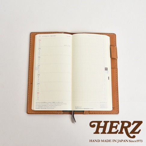 淺棕色皮革製成的旅行記事本書套，打開展示內頁，左頁為日曆格式，右頁為方格筆記頁，書套邊緣有縫線裝飾，下方有品牌標誌。