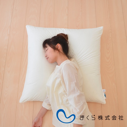 一位女性側臥於一張純白色、柔軟的方枕上，枕頭置於淺色木質地板上。女性身穿米白色長袖上衣，頭髮盤起。枕頭一角貼有藍色標籤。