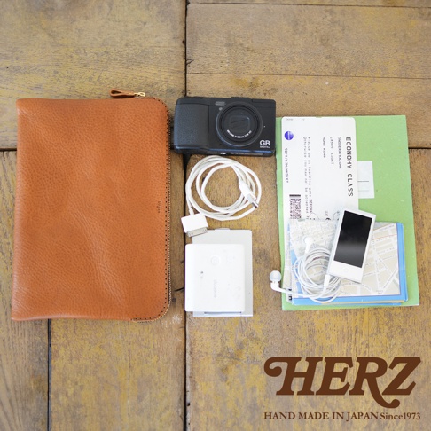 職人手作皮革Organ雙拉鍊收納袋，呈淺棕色，皮革材質，表面有自然紋理。袋子旁有黑色數位相機、白色充電線、白色行動電源、白色耳機、 iPod touch 和一張綠色機票。