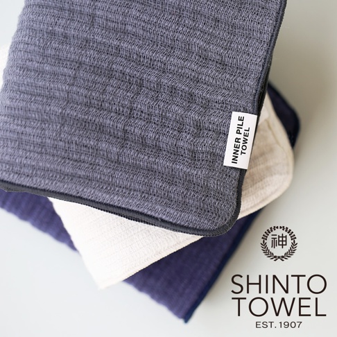 三條疊放的 INNER PILE 雙面紗布毛巾，頂部為深灰色，中間為米白色，底部為海軍藍。毛巾表面織紋清晰，材質輕薄。