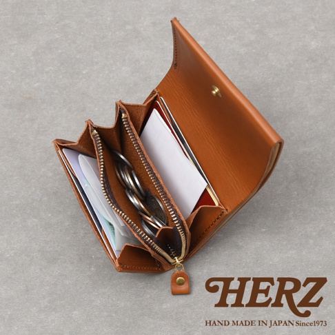 由日本HERZ職人手作的皮革雙折迷你短夾，選用義大利Minerva Liscio天然皮革製成。錢包採暖棕色調，內部結構設計精密，中央設有帶拉鍊的零錢收納袋，兩側皆有寬敞的隔層，方便分類存放卡片、紙鈔與單據，整體展現極簡俐落的工藝美感。