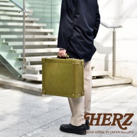 身著海軍藍西裝外套的男士，手提一個橄欖綠色職人手作皮革英倫風便攜手提行李箱，箱子角落有金色金屬加固，搭配米色長褲和黑色皮鞋，背景為現代階梯。