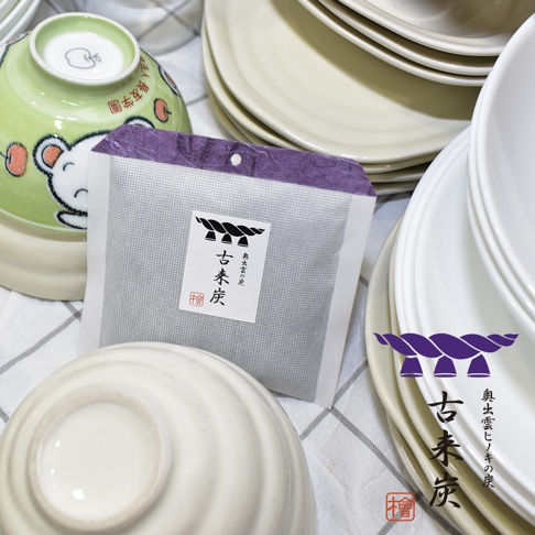 古來炭迷你調濕木炭包，白色包裝，紫色頂部，內含黑色木炭顆粒，印有中文「古來炭」字樣及品牌標誌。背景為白色陶瓷碗碟與綠色卡通碗。