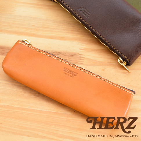 職人手作皮革經典收納筆袋，展現兩種顏色：淺棕色與深棕色。兩款筆袋均採用真皮材質，搭配金色拉鍊與手工縫線，設計簡約。