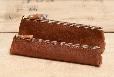 兩個棕色皮革製的三角收納筆袋，配有金色拉鍊和皮革拉環，放置在木質背景上。