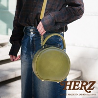 日本HERZ品牌，職人手作皮革兩用圓形包，綠色皮革搭配牛仔裙，包上印有HERZ品牌字樣。