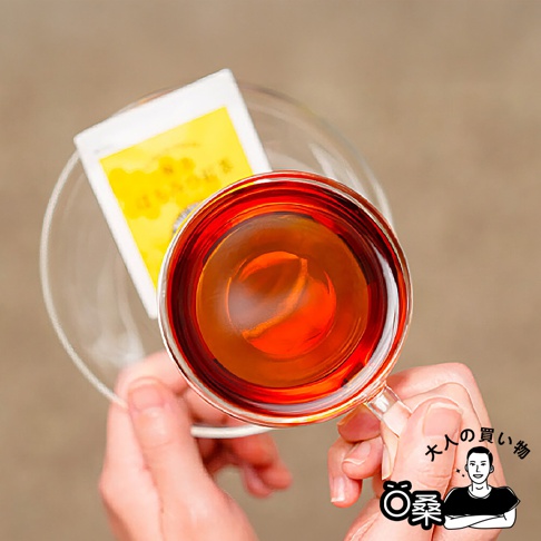 俯視視角下，手持一杯琥珀色紅茶，杯旁放著一個印有日文的白色茶包，背景為米色。