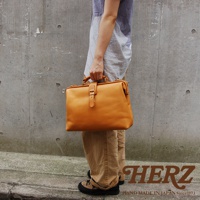 職人手作皮革軟式杜勒斯包，採用天然皮革材質，呈現溫潤的焦糖色。包款設計經典，配有金屬扣環及提把，由一側展示。